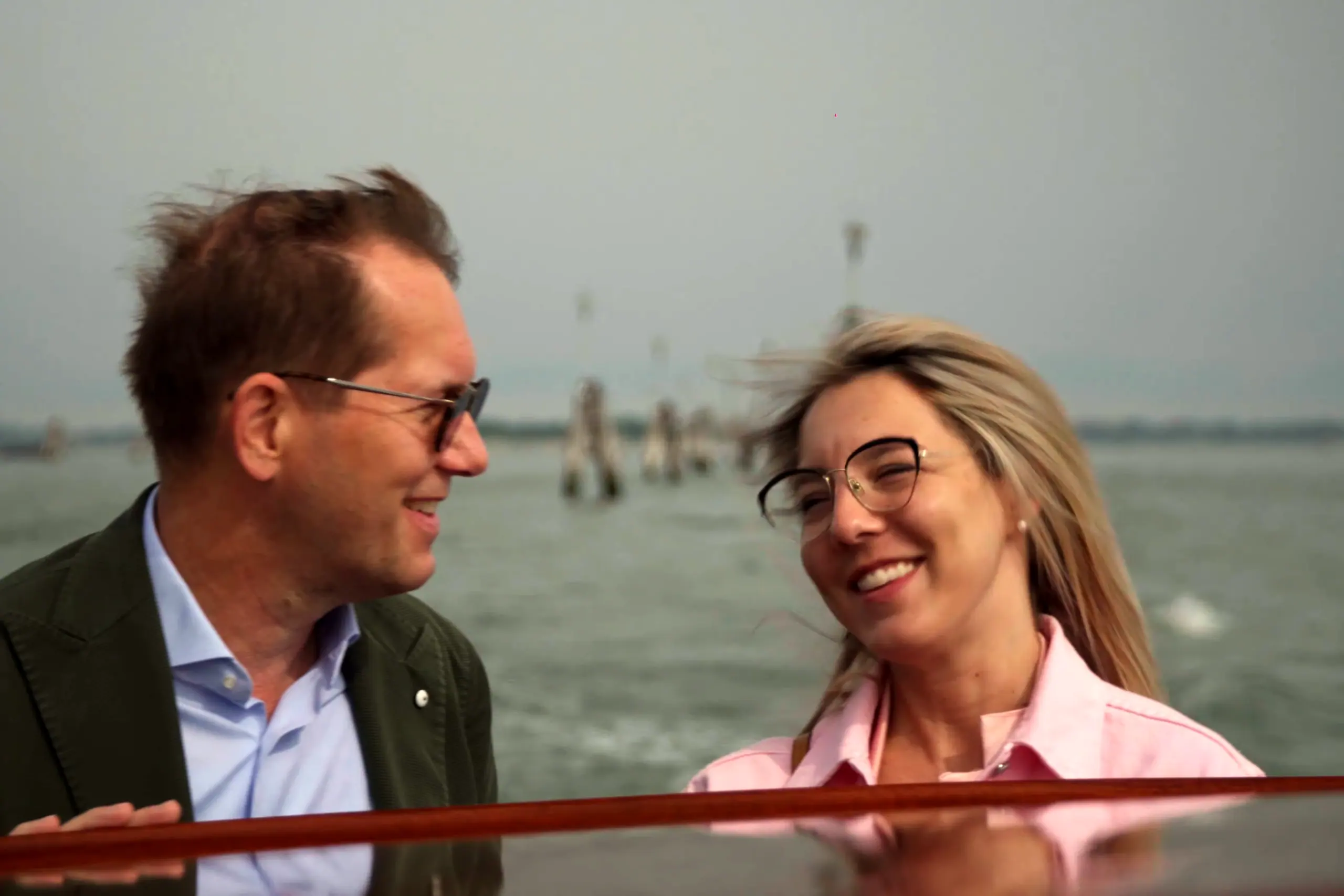 Il Dott. Ronald Brouwers sorride in barca nella laguna, accanto a una donna in camicia rosa; dietro, le bricole
