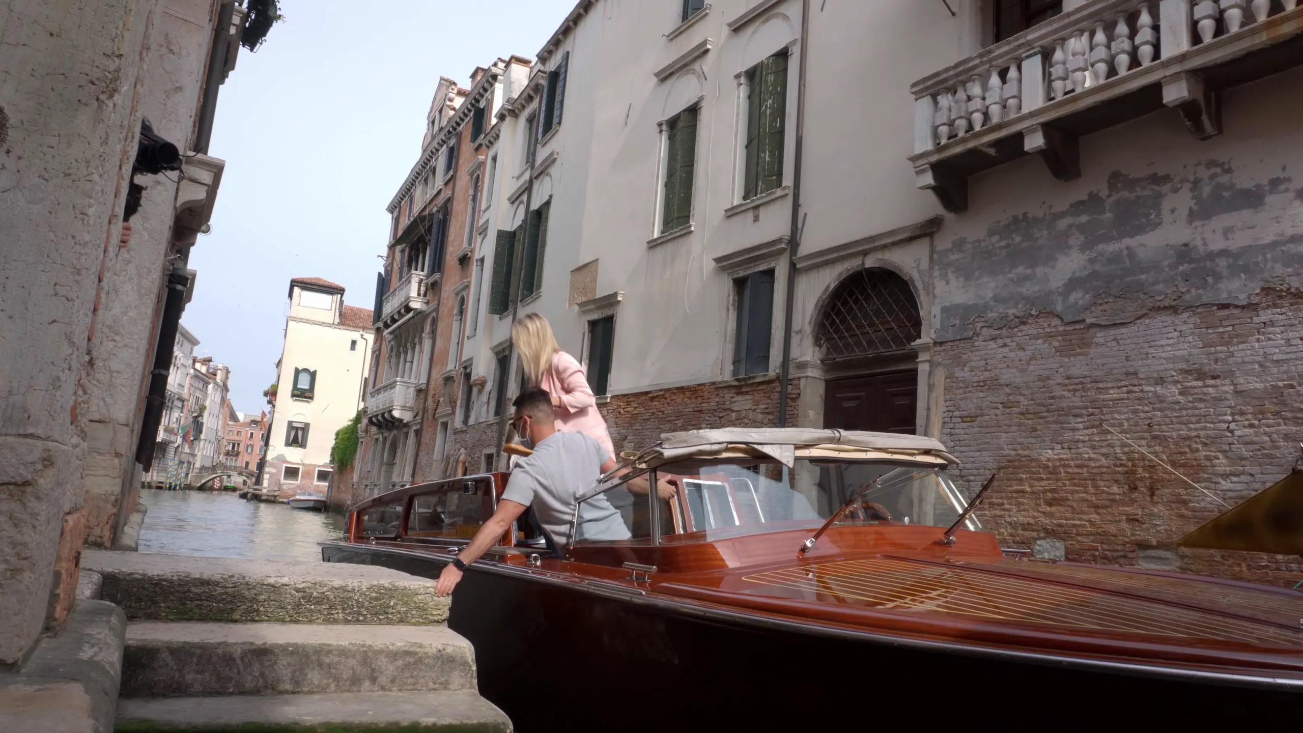 Una barca di legno ormeggiata ai gradini di una riva, in un rio di Venezia fra i palazzi; una passeggera sta per scendere